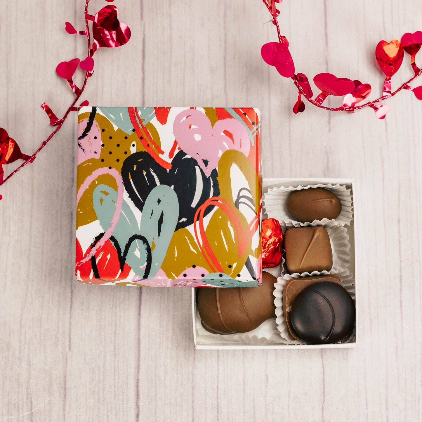a square box with colorful fluttering hearts on it is filled with 3 oz. of assorted chocolates