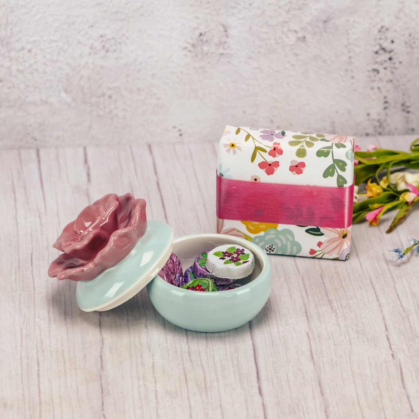 A ceramic teal trinket dish with a rose colored flower lid is packaged with some sweet treats