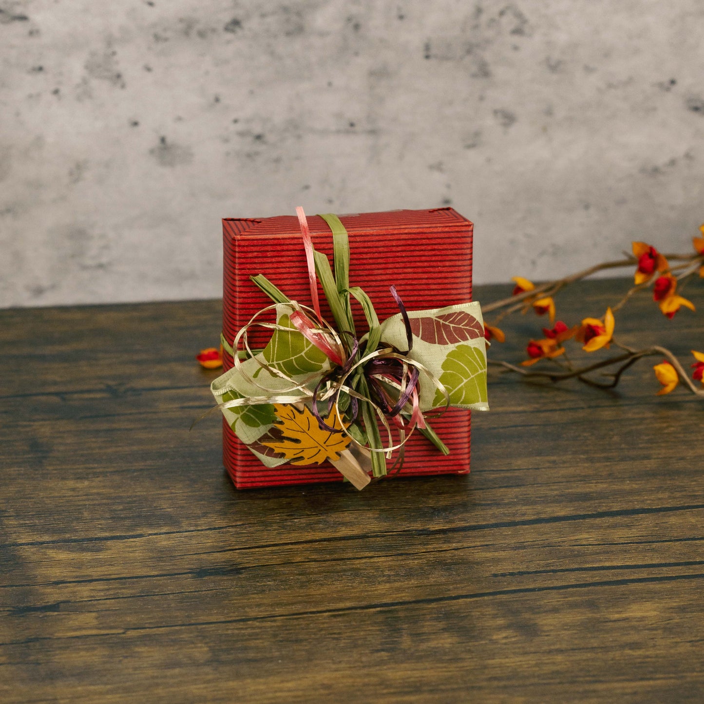 a sampler box of assorted chocolates is wrapped in fall maroon wrap and topped with a festive handmade bow and a leaf wooden clip.