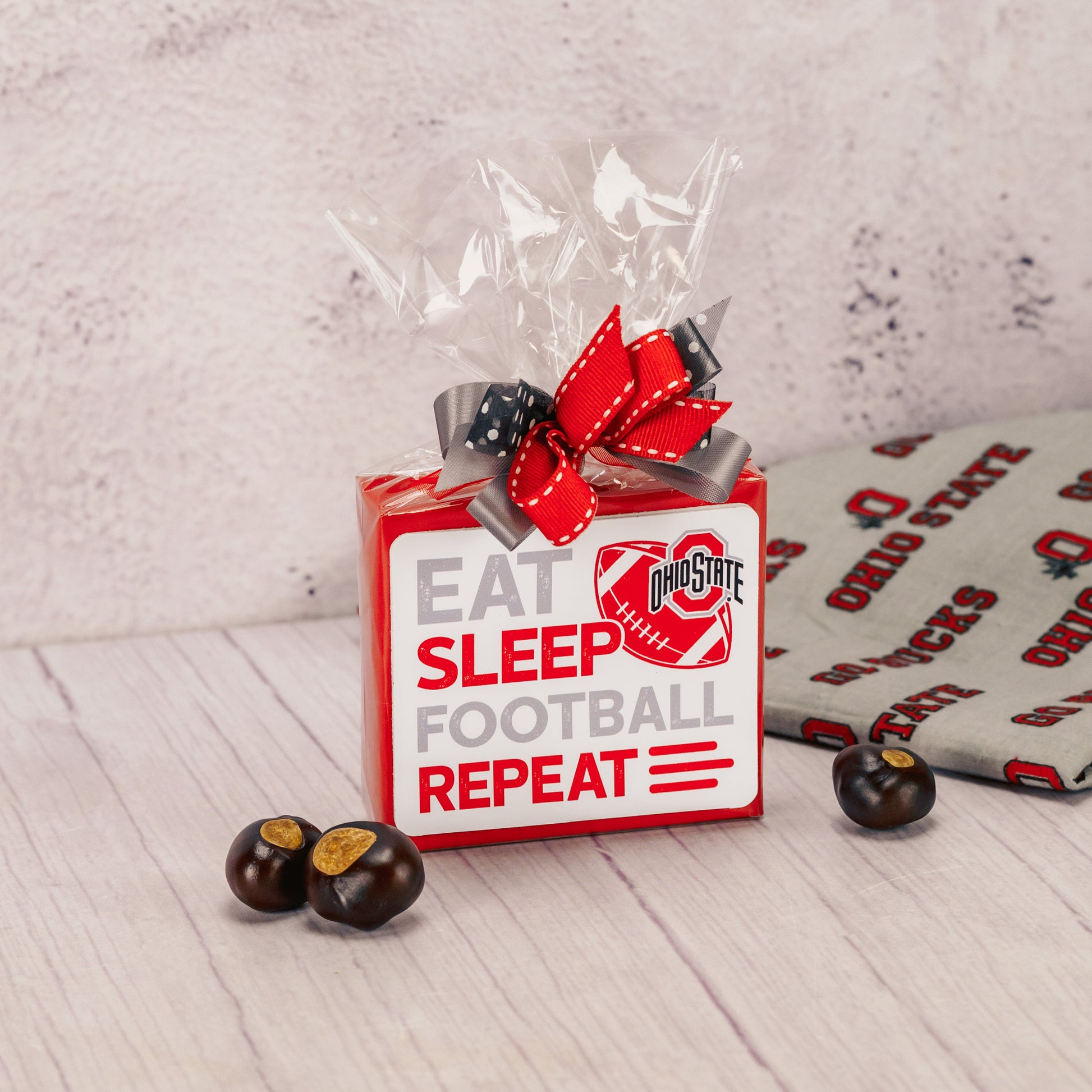 a sampler box of assorted chocolates is wrapped and topped with an Ohio State football sticker that reads 'eat, sleep, football, repeat'.