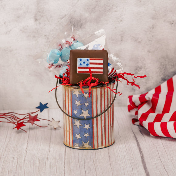a patriotic graham cracker, raspberry taffy and milk chocolate american foil hearts are stuffed inside a metal pail decorated with stars and stripes.