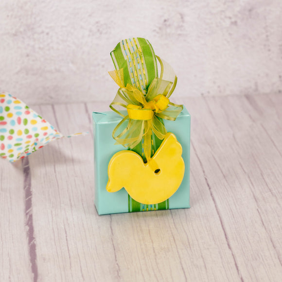 a sampler box of assorted chocolates is wrapped and topped with a yellow ceramic duck ornament