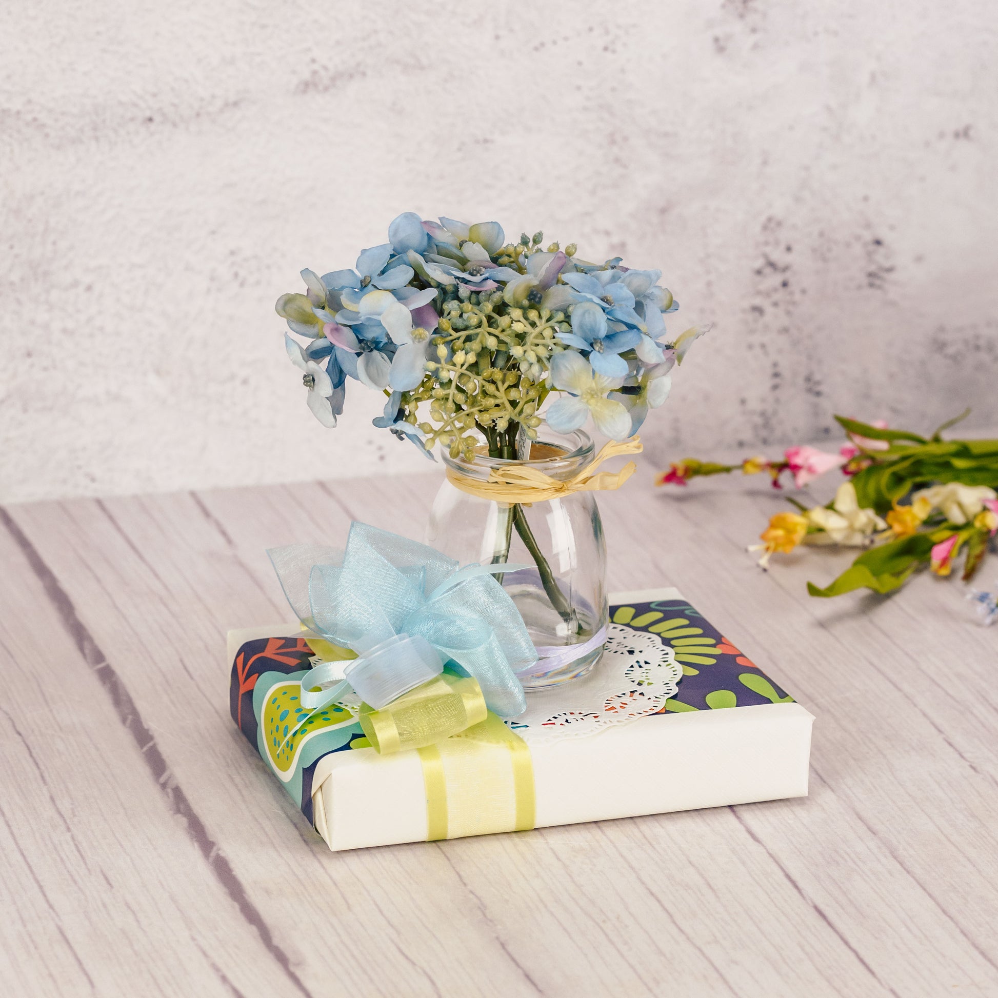 a half pound box of assorted chocolates is wrapped and topped with a glass vase of blue hydrangea sprigs.
