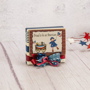 a sampler box of assorted chocolates is wrapped patriotically and topped with a wooden magnet that reads 'Proud to be an American'
