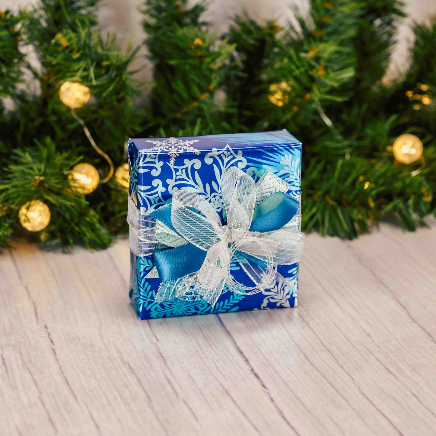 a sampler box of assorted chocolates is wrapped in blue shiny snowflake paper with a handmade bow on top.