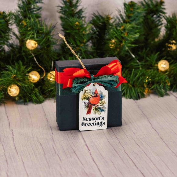 a sampler box of assorted chocolates is wrapped and topped with a flat wooden cardinal ornament that says 'season's greetings'