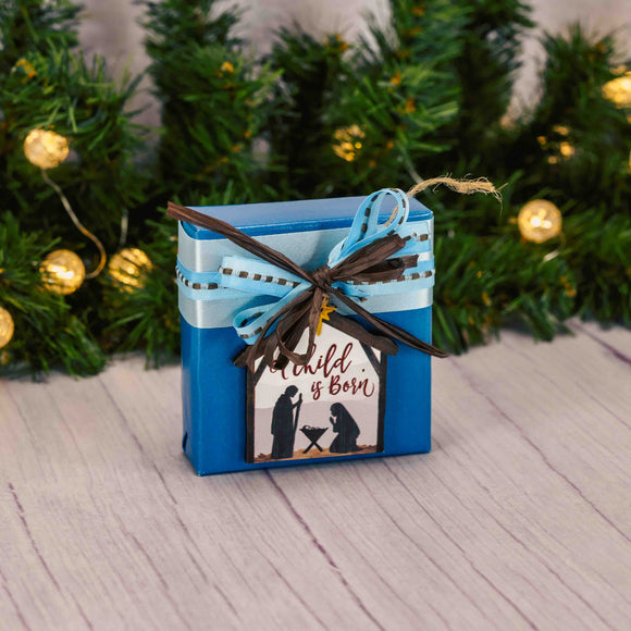 a sampler box of assorted chocolates is wrapped in blue and topped with a flat wooden nativity ornament that reads 'a child is born'