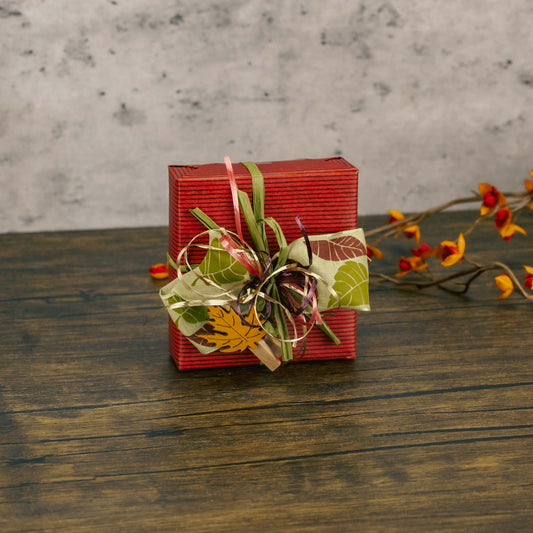 a sampler box of assorted chocolates is wrapped in fall maroon wrap and topped with a festive handmade bow and a leaf wooden clip.