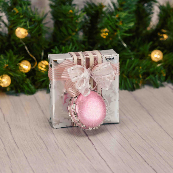 a sampler box of assorted chocolates is wrapped in silver holly paper and topped with a pink embellished macaron ornament