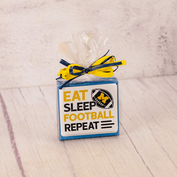a sampler box of assorted chocolates wrapped and topped with a Michigan football sticker that reads 'eat, sleep, football, repeat'