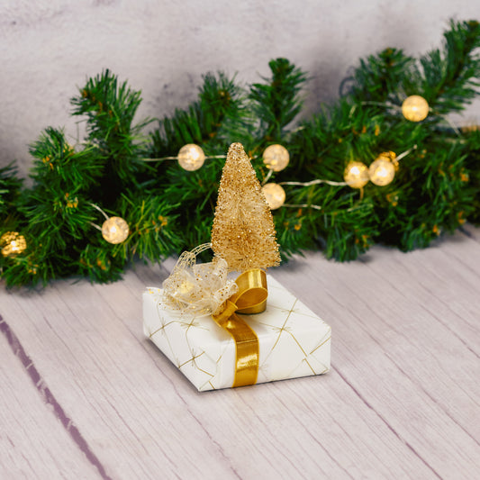 a sampler box of assorted chocolates is wrapped in ivory and gold and topped with an ivory bristle tree with gold glitter