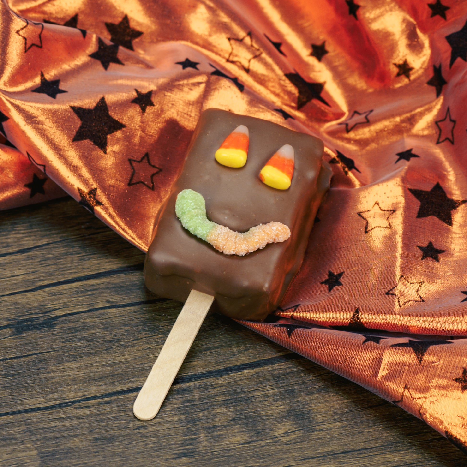 A rectangle Rice Krispie Treat on a stick covered in milk chocolate with a sour neon worm for a mouth and candy corn for eyes.