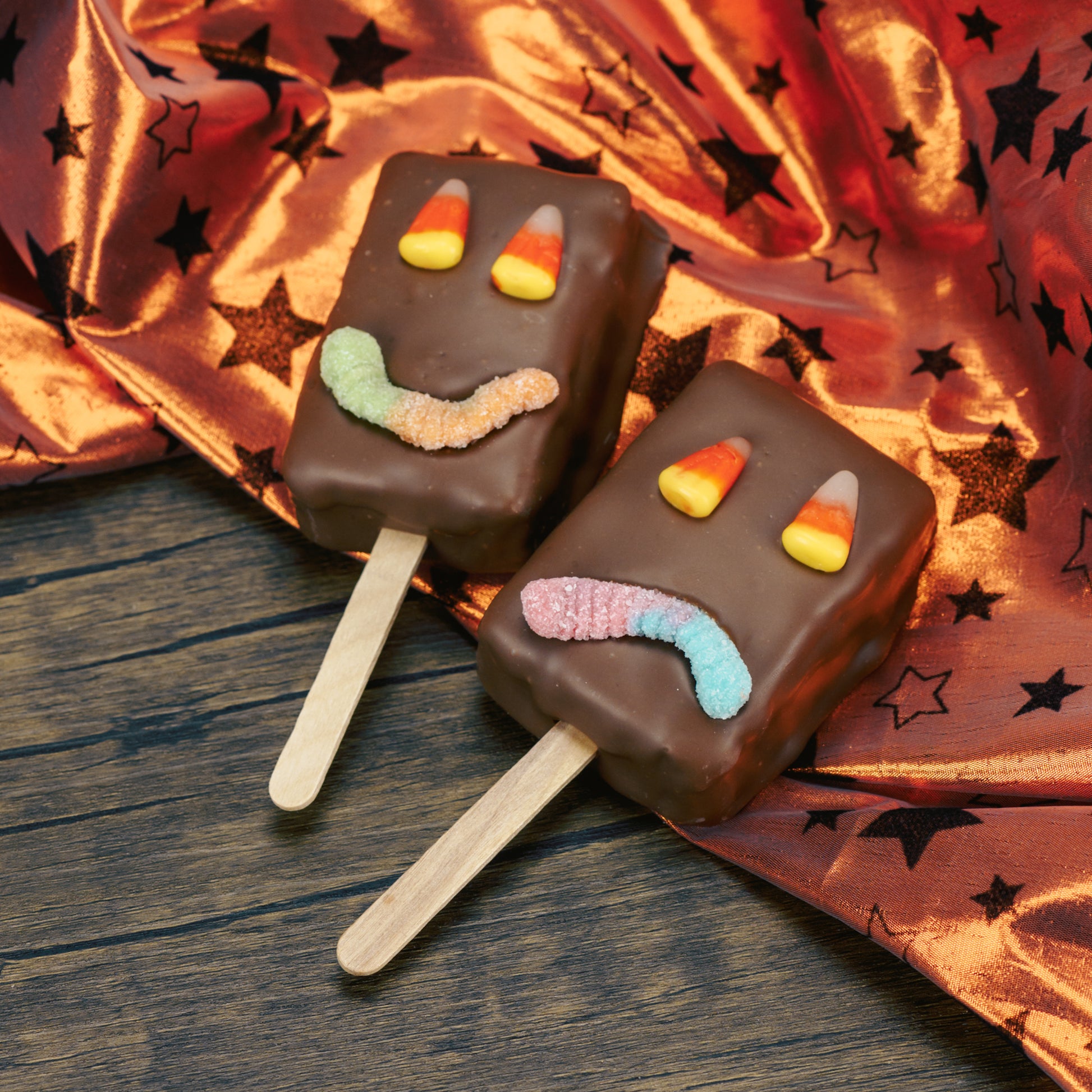 A rectangle Rice Krispie Treat on a stick covered in milk chocolate with a sour neon worm for a mouth and candy corn for eyes.